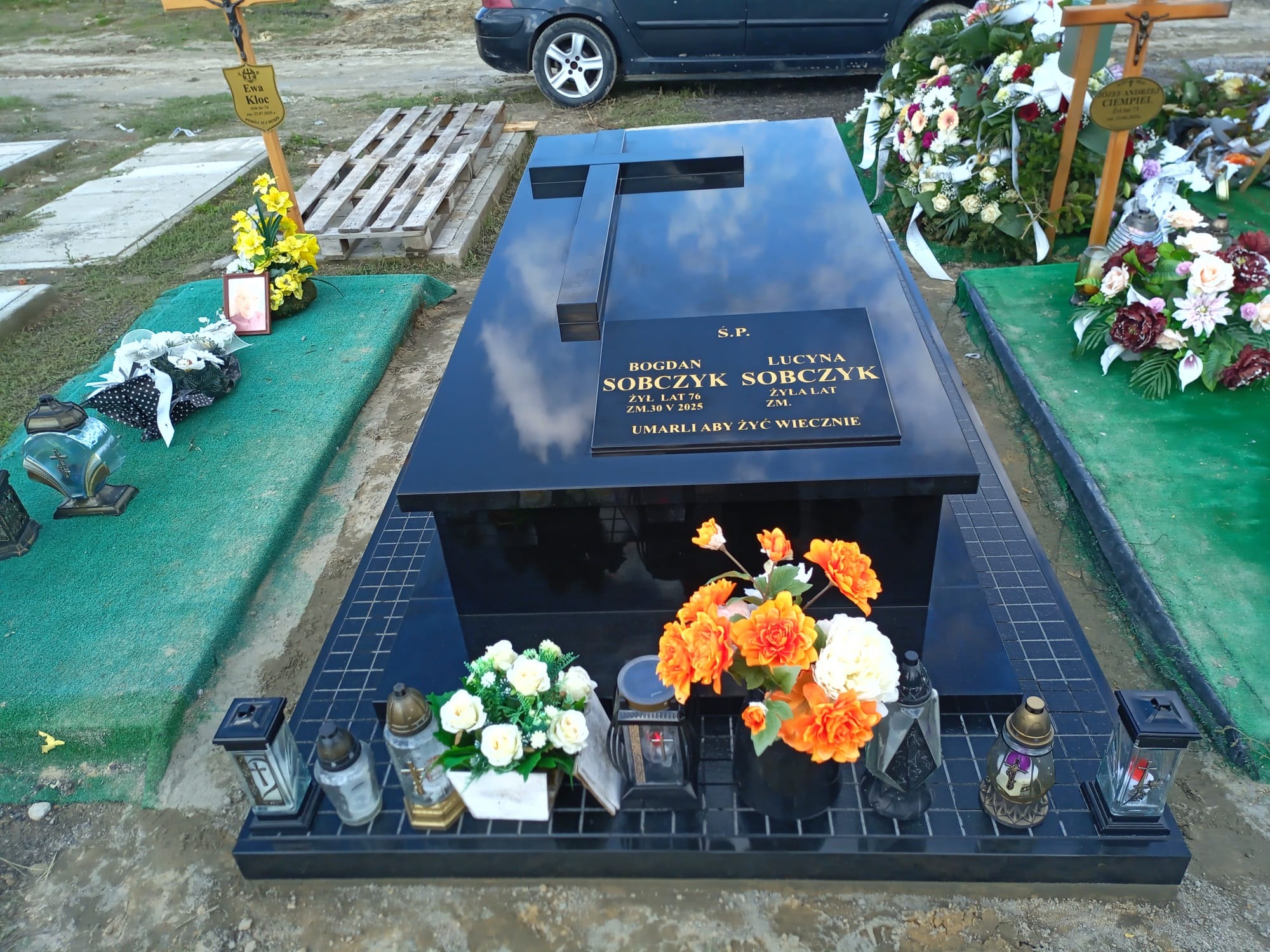 Czarny, nowoczesny nagrobek z krzyżem i inskrypcją Sobczyk, otoczony kwiatami i lampionami na cmentarzu. Błyszcząca powierzchnia odbija niebo.