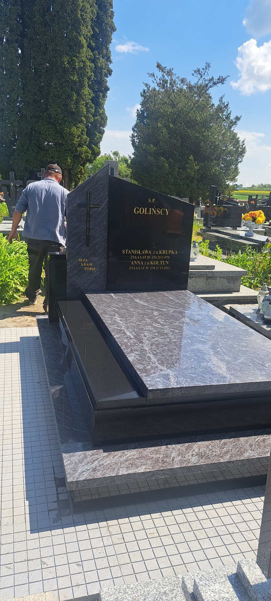 Czarny, polerowany nagrobek z szarego granitu na cmentarzu, z inskrypcjami i krzyżem. Mężczyzna w tle. Słoneczny dzień, widok na inne groby i zieleń.