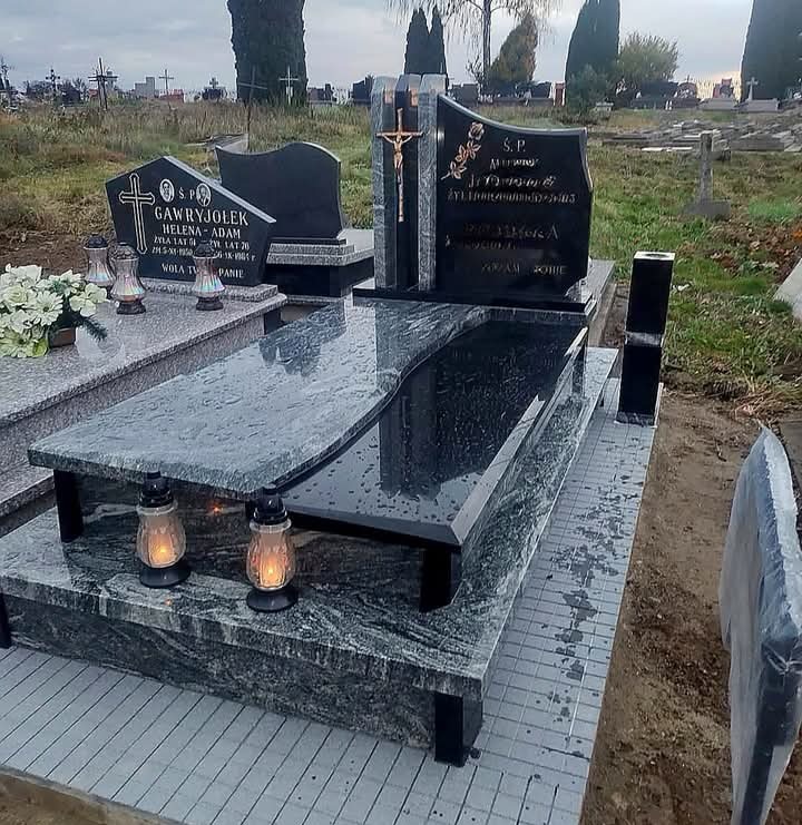 Czarny, polerowany nagrobek z krzyżem i inskrypcjami na cmentarzu, z zapalonymi lampionami. Widok z boku, pochmurne niebo w tle.