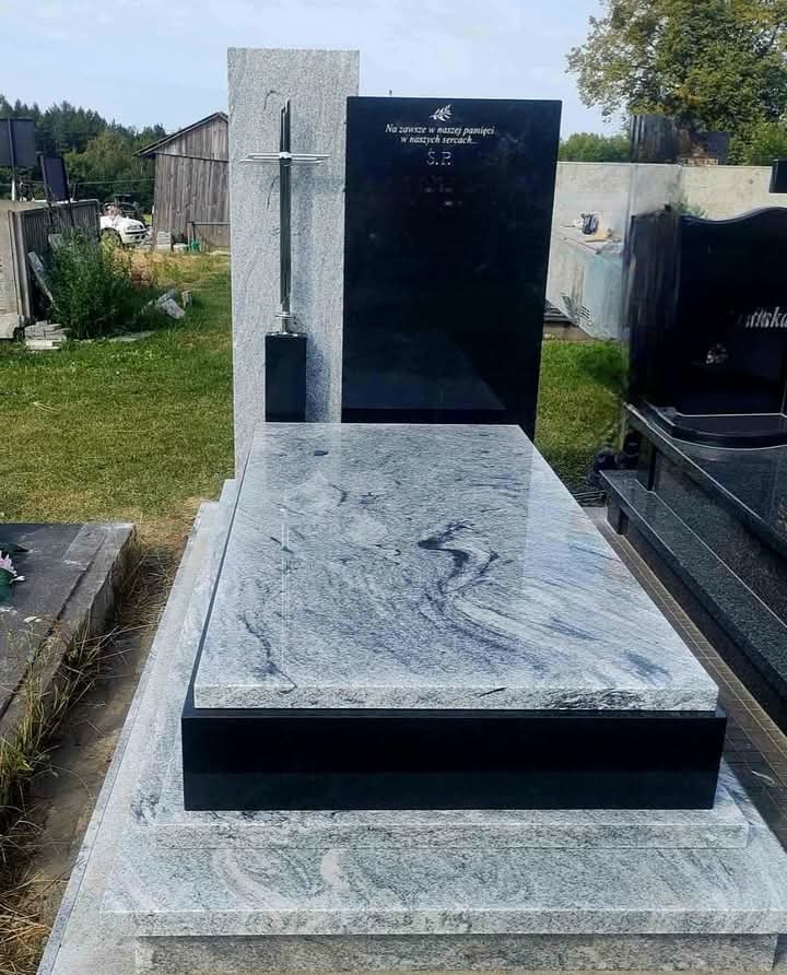 Granitowy nagrobek z czarną tablicą inskrypcyjną i srebrnym krzyżem, na tle zieleni. Jasnoszary kamień z wyraźnym wzorem żyłkowania.