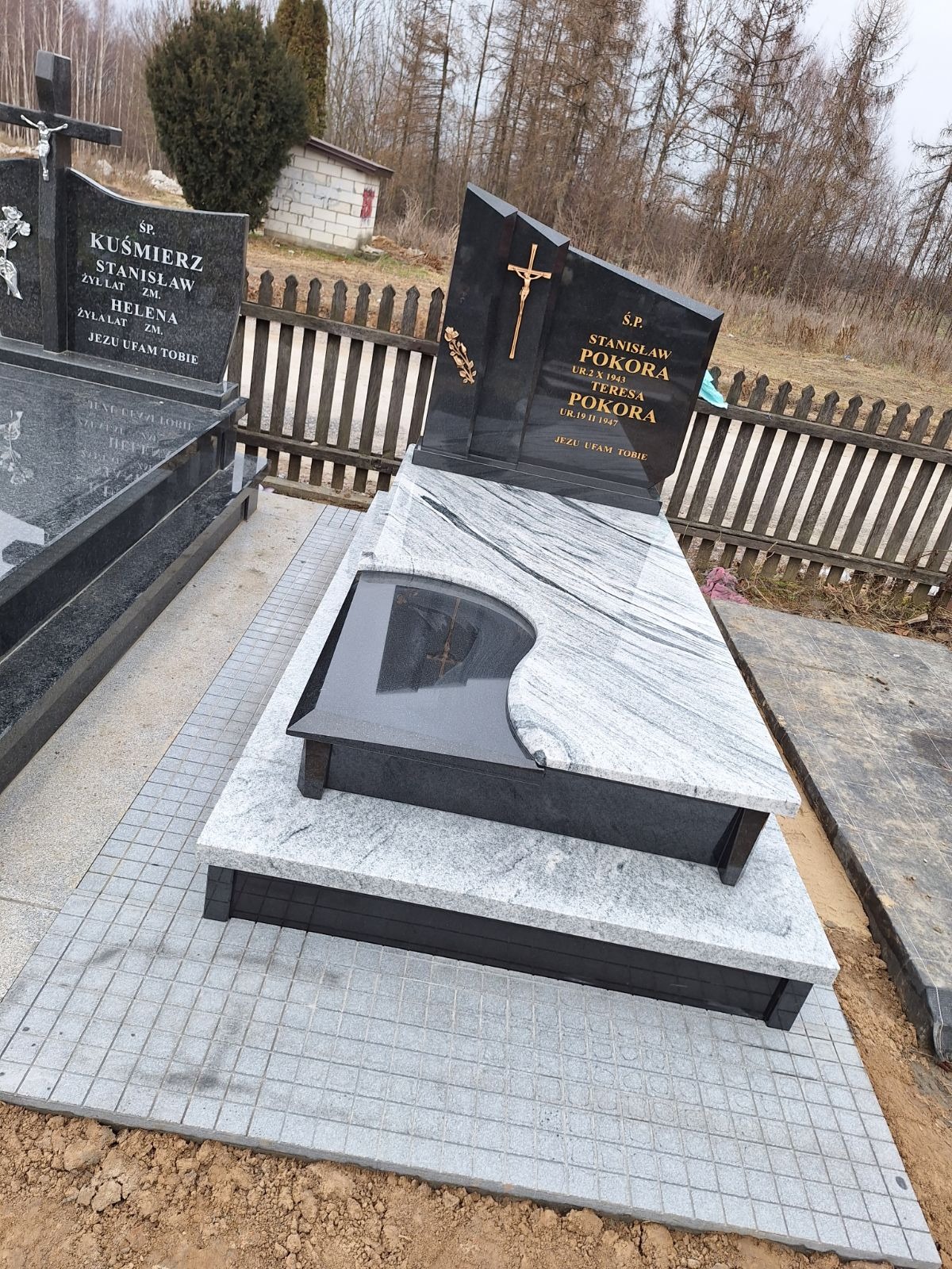 Nowoczesny nagrobek z jasnego granitu z ciemnymi elementami, zdobiony złotym krzyżem i inskrypcjami. Widoczne są sąsiednie nagrobki i drewniane ogrodzenie.