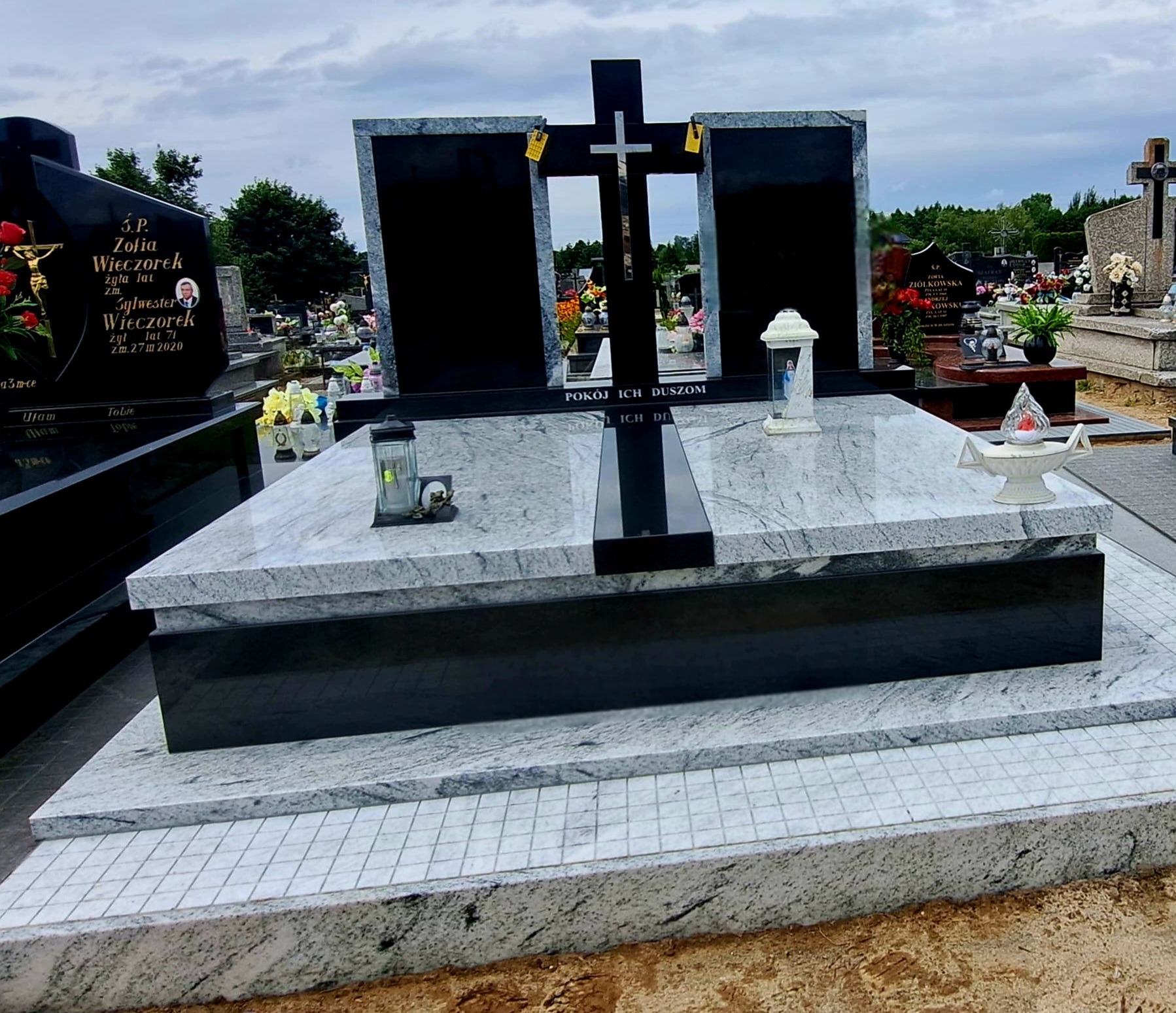 Dwukolorowy nagrobek z krzyżem na cmentarzu, wykonany z jasnego i ciemnego kamienia. Widoczne znicze i napisy. Tło stanowią inne nagrobki i drzewa.