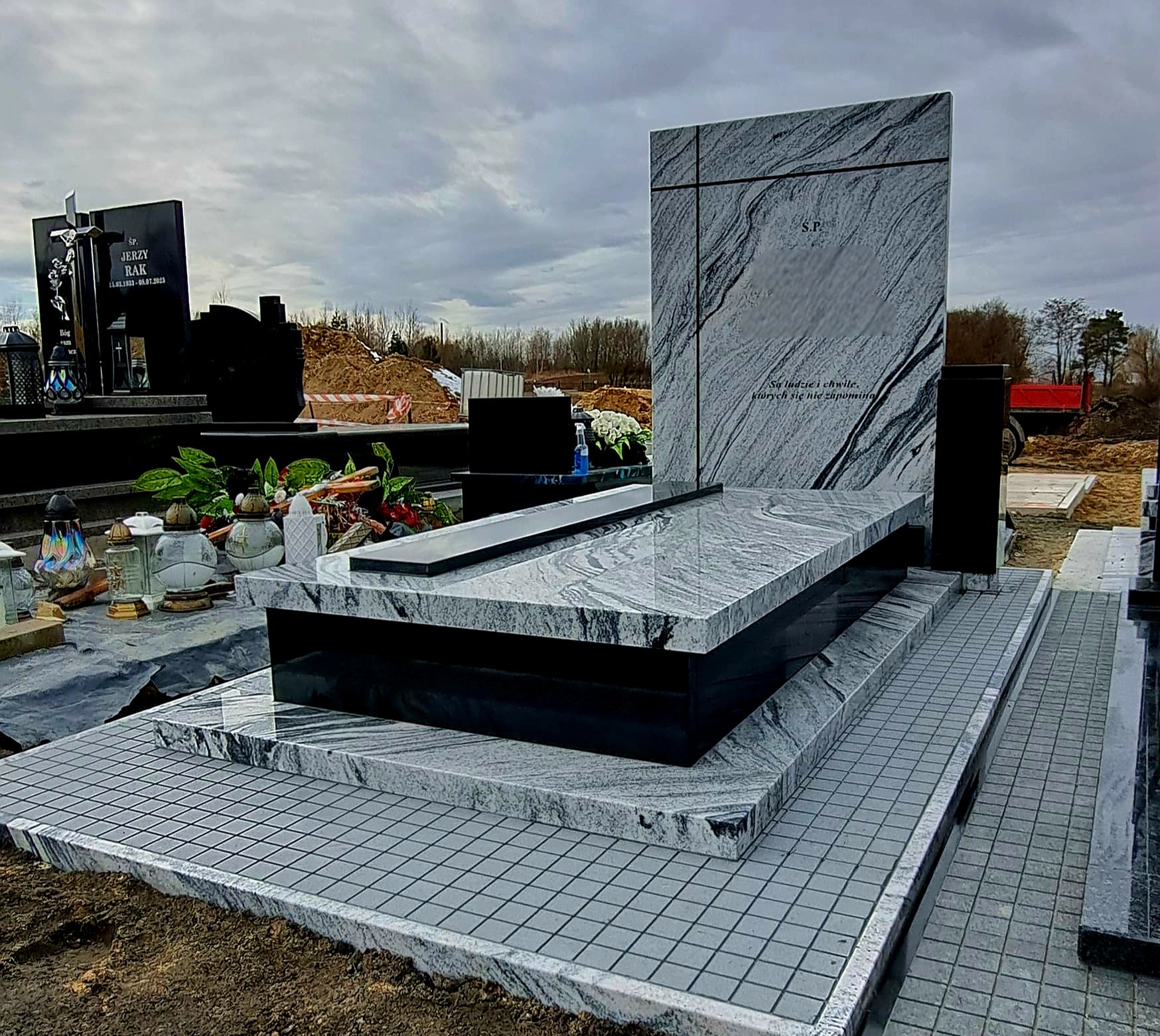 Nowoczesny nagrobek z szarego granitu z czarnymi elementami, na cmentarzu. Widoczne kwiaty i znicze. Tło: pochmurne niebo i fragmenty innych nagrobków.