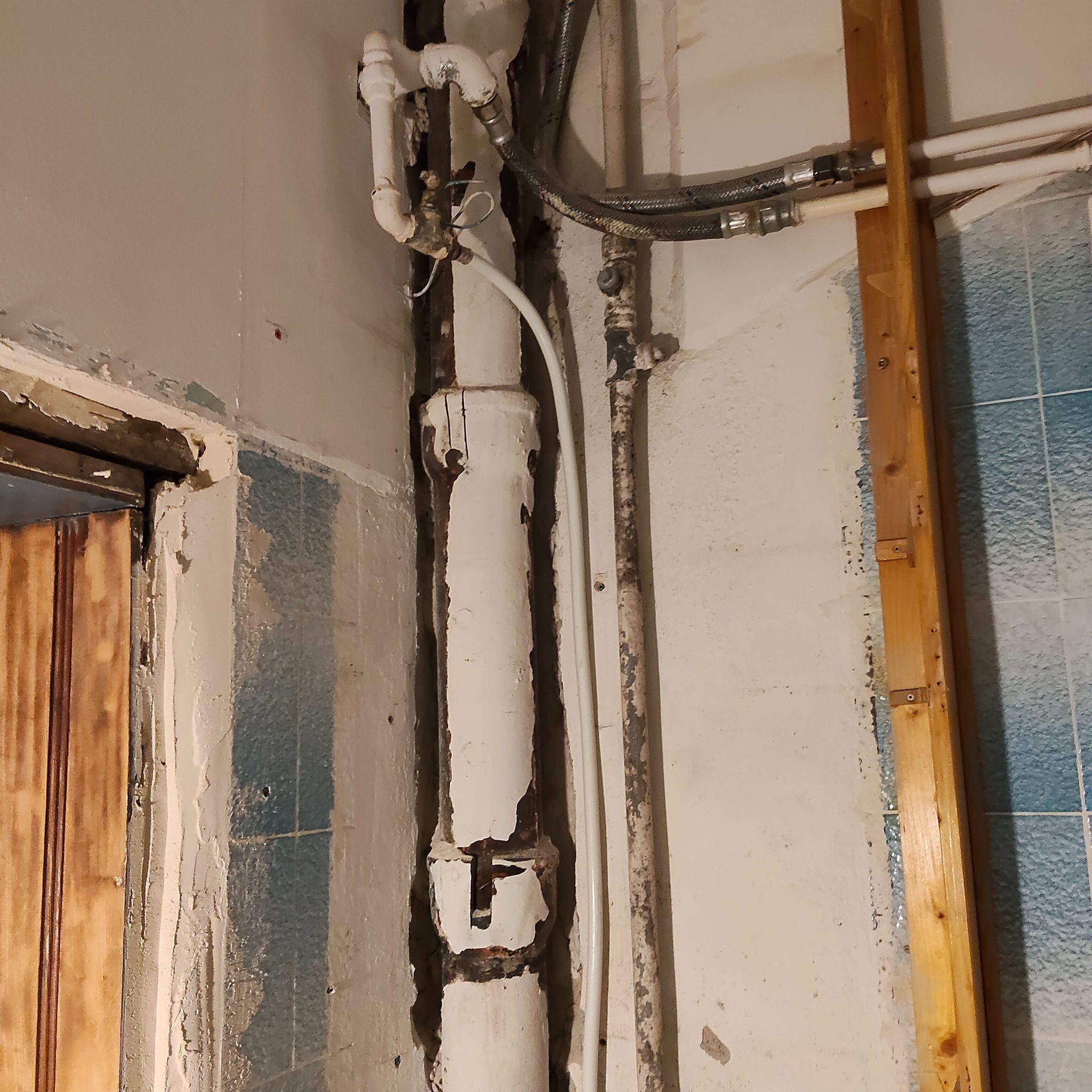 Zniszczona ściana z rurami w łazience przed remontem. Widoczne odpryski farby, stare płytki i elementy instalacji wodnej. Fragment drewnianej ramy okiennej.