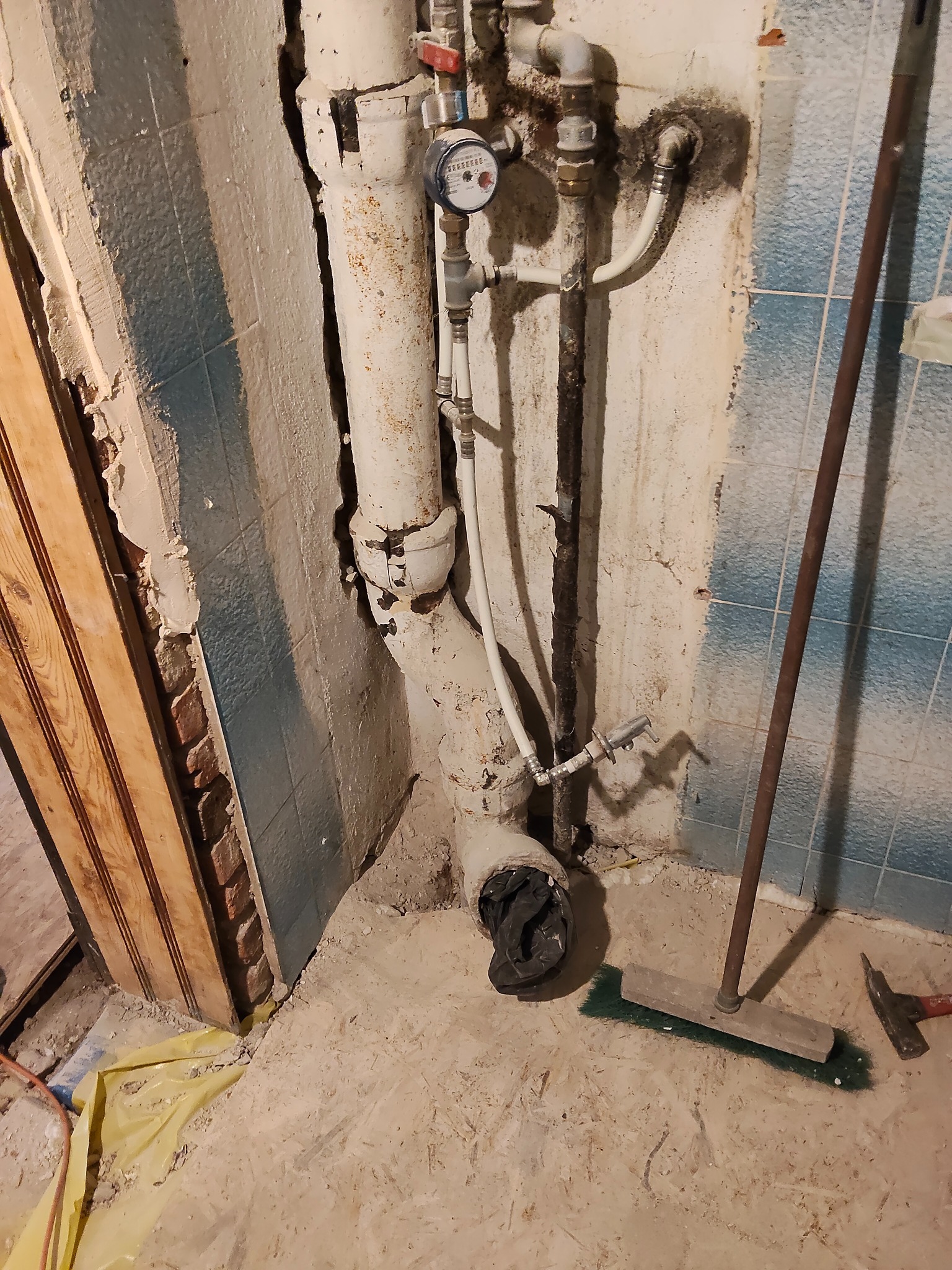 Niefotogeniczne ujęcie rur i fragmentu ściany w trakcie remontu. Widoczne stare płytki, fragment odsłoniętej cegły i narzędzia na podłodze.