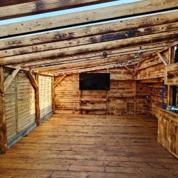 Roman Zhukov - Zadaszona konstrukcja z drewna o unikalnym wzorze słojów, z podłogą z desek i zamontowanym telewizorem. Widoczne oświetlenie i elementy wyposażenia.
