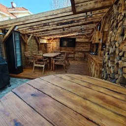 Roman Zhukov - Drewniana altana z wyposażeniem: stół, krzesła, lodówka i telewizor. Ściana z ułożonego drewna opałowego. Widok z perspektywy stołu na pierwszym planie.