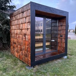 Roman Zhukov - Nowoczesna, drewniana altana w kształcie sześcianu, pokryta gontem, z przeszklonymi drzwiami, na tle zieleni i budynku biurowego. Minimalistyczny design.