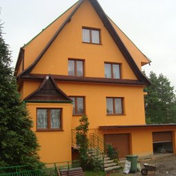 Piotr Ficek ZAKŁAD USŁUGOWO-REMONTOWO-BUDOWLANY - Dwupiętrowy dom z pomarańczową elewacją i brązowymi akcentami dachu, widoczny z zewnątrz w pochmurny dzień. Obok domu rośnie duża choinka.