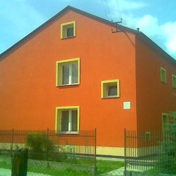 Piotr Ficek ZAKŁAD USŁUGOWO-REMONTOWO-BUDOWLANY - Pomarańczowa elewacja domu z żółtymi obramowaniami okien, widoczna zza metalowego ogrodzenia. Dom dwukondygnacyjny z oknami o różnych rozmiarach.