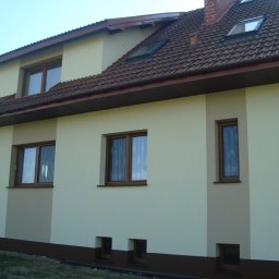 Piotr Ficek ZAKŁAD USŁUGOWO-REMONTOWO-BUDOWLANY - Dwukondygnacyjny dom z beżową elewacją, brązowymi oknami i dachem z czerwonej dachówki. Fragment elewacji w odcieniu brązu. Widok z poziomu gruntu, słoneczny dzień.