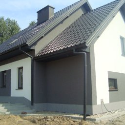 Piotr Ficek ZAKŁAD USŁUGOWO-REMONTOWO-BUDOWLANY - Dwukolorowy dom jednorodzinny z ciemną dachówką i szarą elewacją, z widocznymi schodami wejściowymi i fragmentem ogrodu. Ujęcie z boku, w słoneczny dzień.