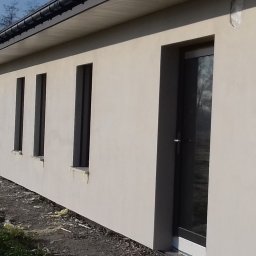 Piotr Ficek ZAKŁAD USŁUGOWO-REMONTOWO-BUDOWLANY - Jasna elewacja domu jednorodzinnego z ciemnymi oknami i drzwiami. Widoczne ślady prac wykończeniowych wokół budynku. Ujęcie z boku, w tle inny budynek.