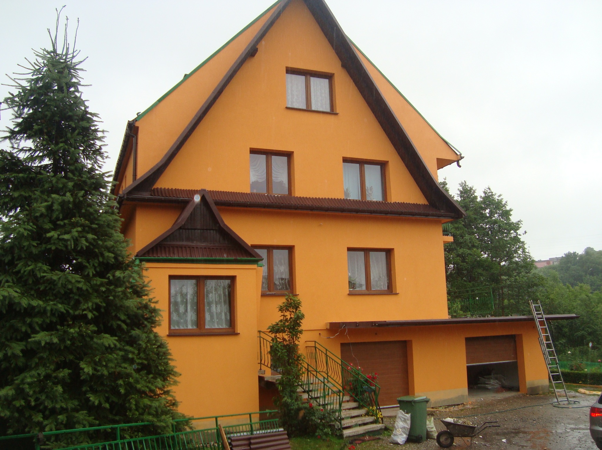 Dwupiętrowy dom z pomarańczową elewacją i brązowymi akcentami dachu, widoczny z zewnątrz w pochmurny dzień. Obok domu rośnie duża choinka.