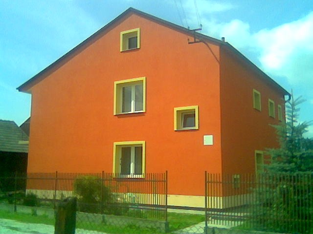 Pomarańczowa elewacja domu z żółtymi obramowaniami okien, widoczna zza metalowego ogrodzenia. Dom dwukondygnacyjny z oknami o różnych rozmiarach.