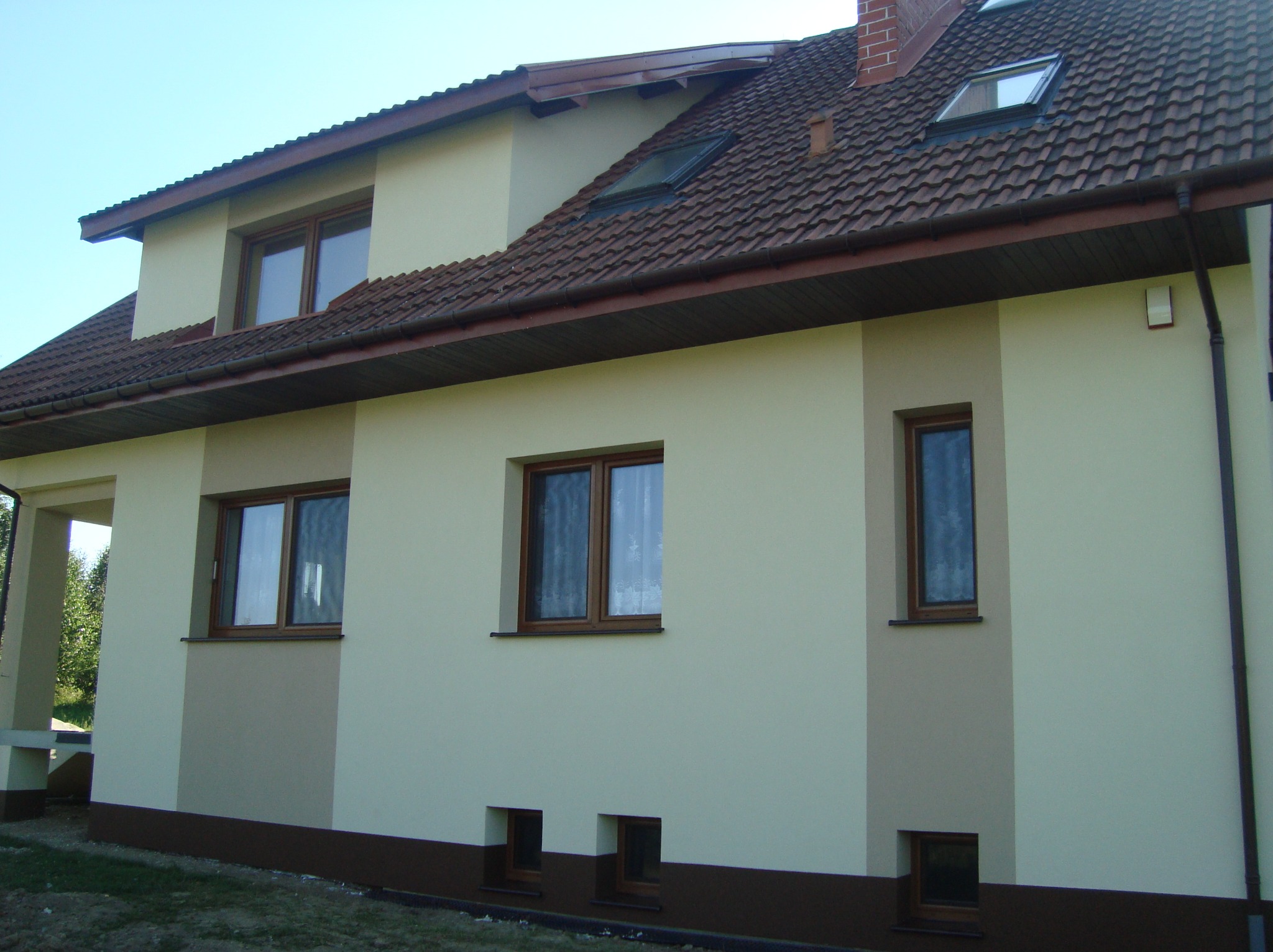 Dwukondygnacyjny dom z beżową elewacją, brązowymi oknami i dachem z czerwonej dachówki. Fragment elewacji w odcieniu brązu. Widok z poziomu gruntu, słoneczny dzień.