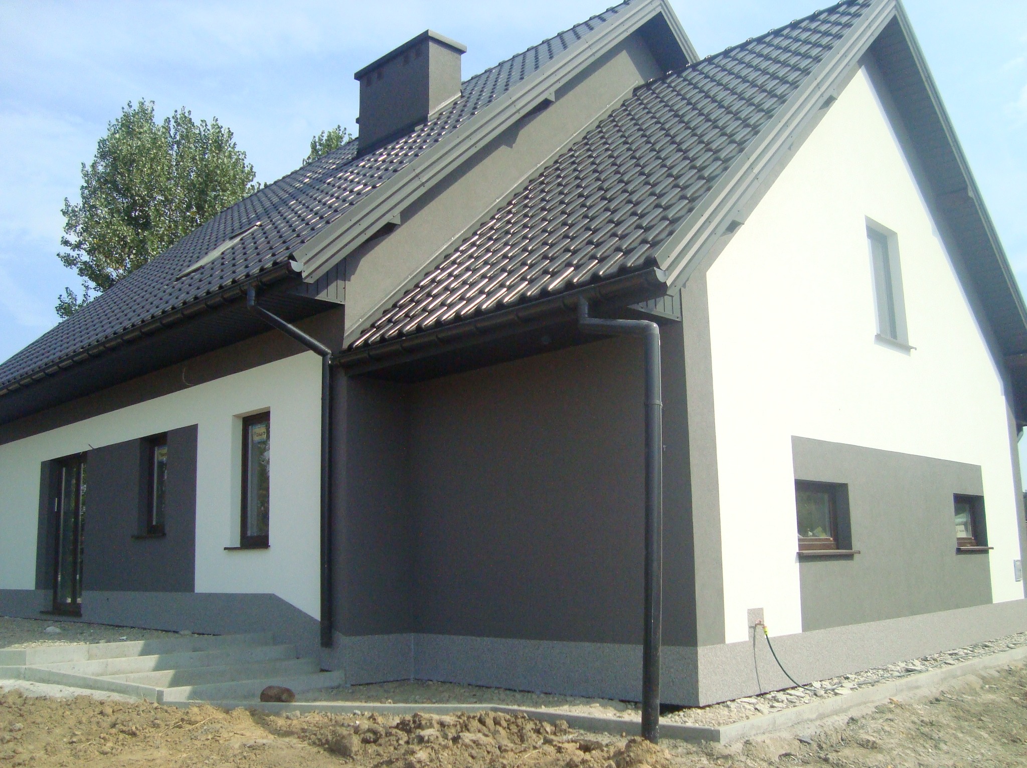 Dwukolorowy dom jednorodzinny z ciemną dachówką i szarą elewacją, z widocznymi schodami wejściowymi i fragmentem ogrodu. Ujęcie z boku, w słoneczny dzień.