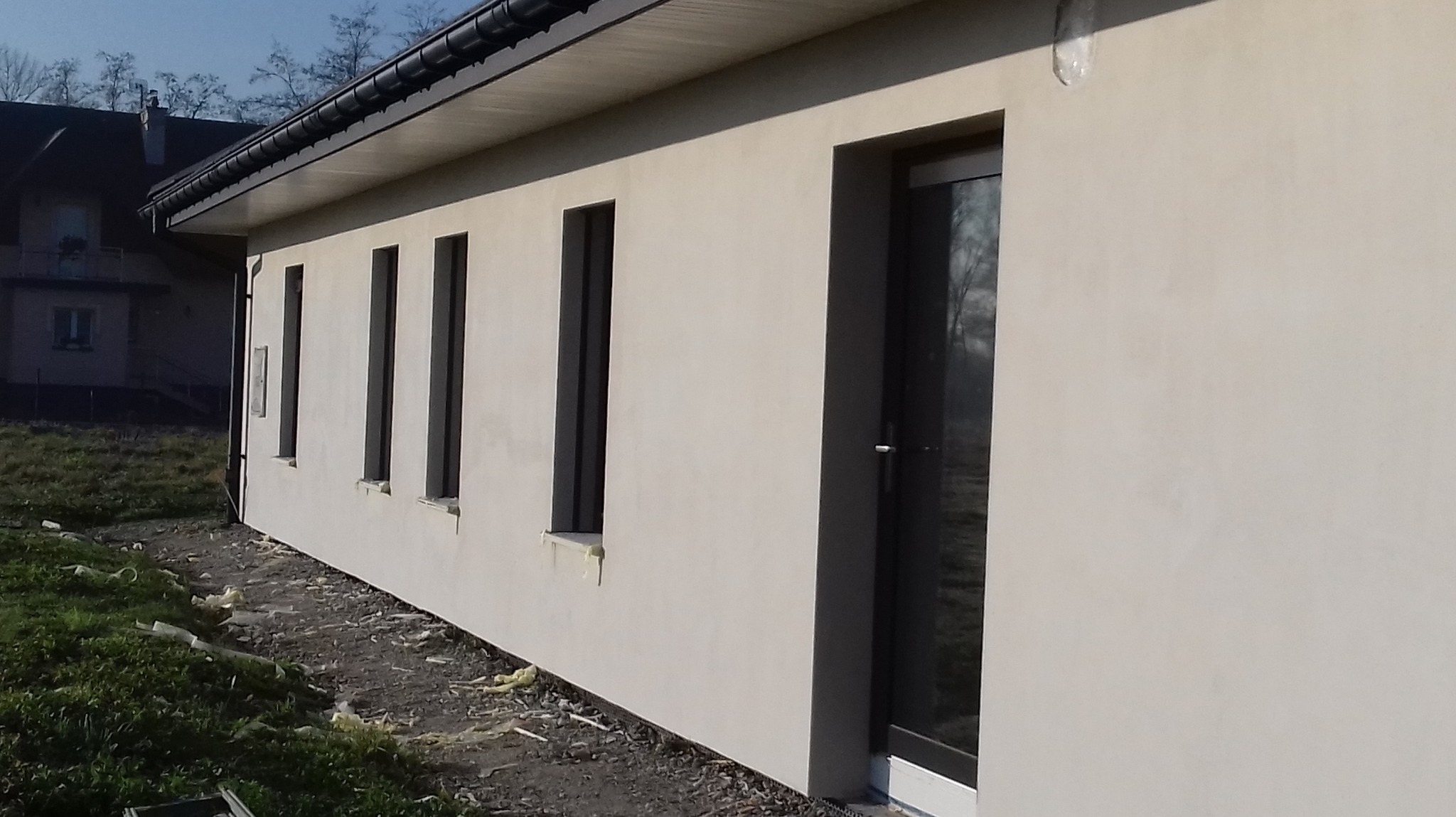 Jasna elewacja domu jednorodzinnego z ciemnymi oknami i drzwiami. Widoczne ślady prac wykończeniowych wokół budynku. Ujęcie z boku, w tle inny budynek.