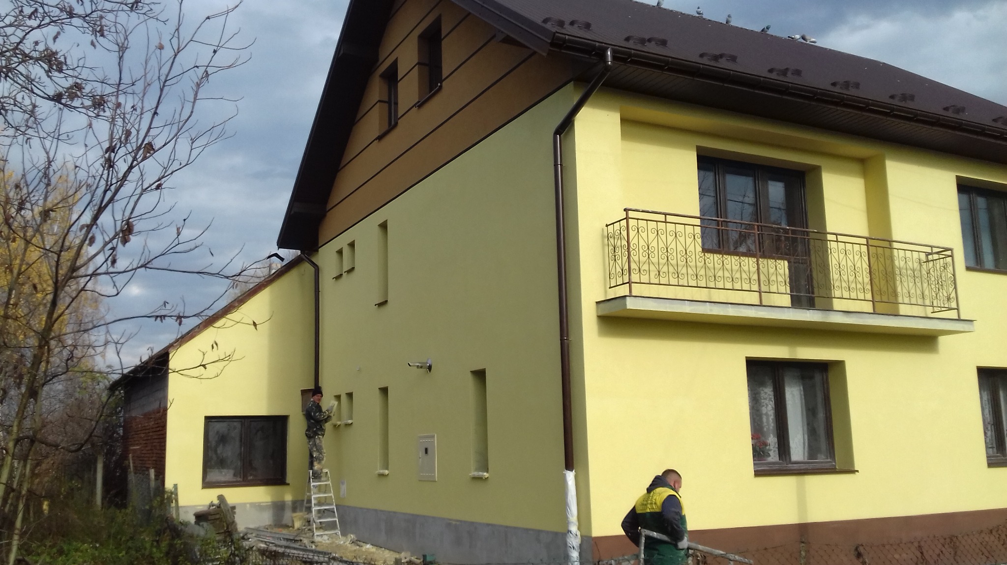 Żółta elewacja domu jednorodzinnego z brązowymi akcentami, pracownicy na drabinie i przy gruncie, pochmurne niebo w tle. Widok z boku.