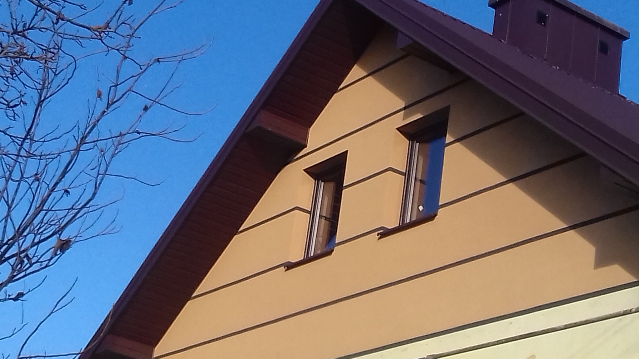 Fragment elewacji domu z dwoma oknami, wykończonej w odcieniach beżu i brązu, z widocznym fragmentem dachu i komina na tle błękitnego nieba i gałęzi drzewa.