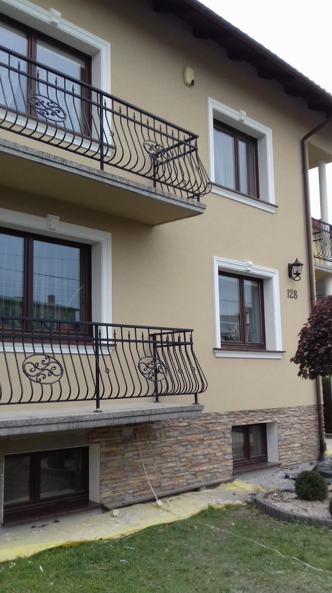 Elewacja domu z beżowym tynkiem, dekoracyjnymi balkonami z kutą balustradą i kamienną podmurówką. Widoczne okna z białymi obramowaniami i numer domu.
