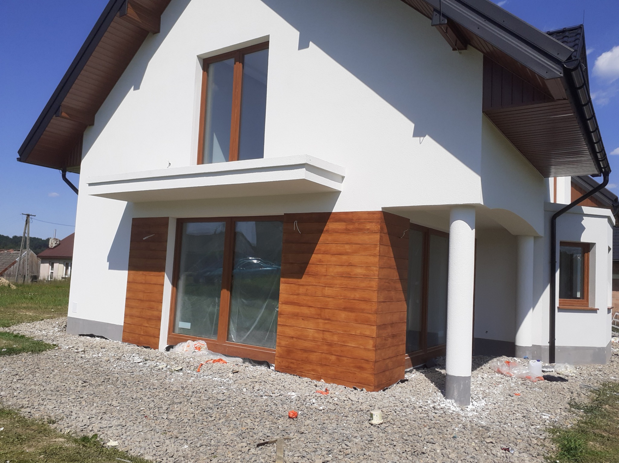 Nowoczesny dom z białą elewacją i drewnianymi panelami. Widoczne okna z brązowymi ramami i grafitowy dach. Otoczenie domu to kamienisty teren.