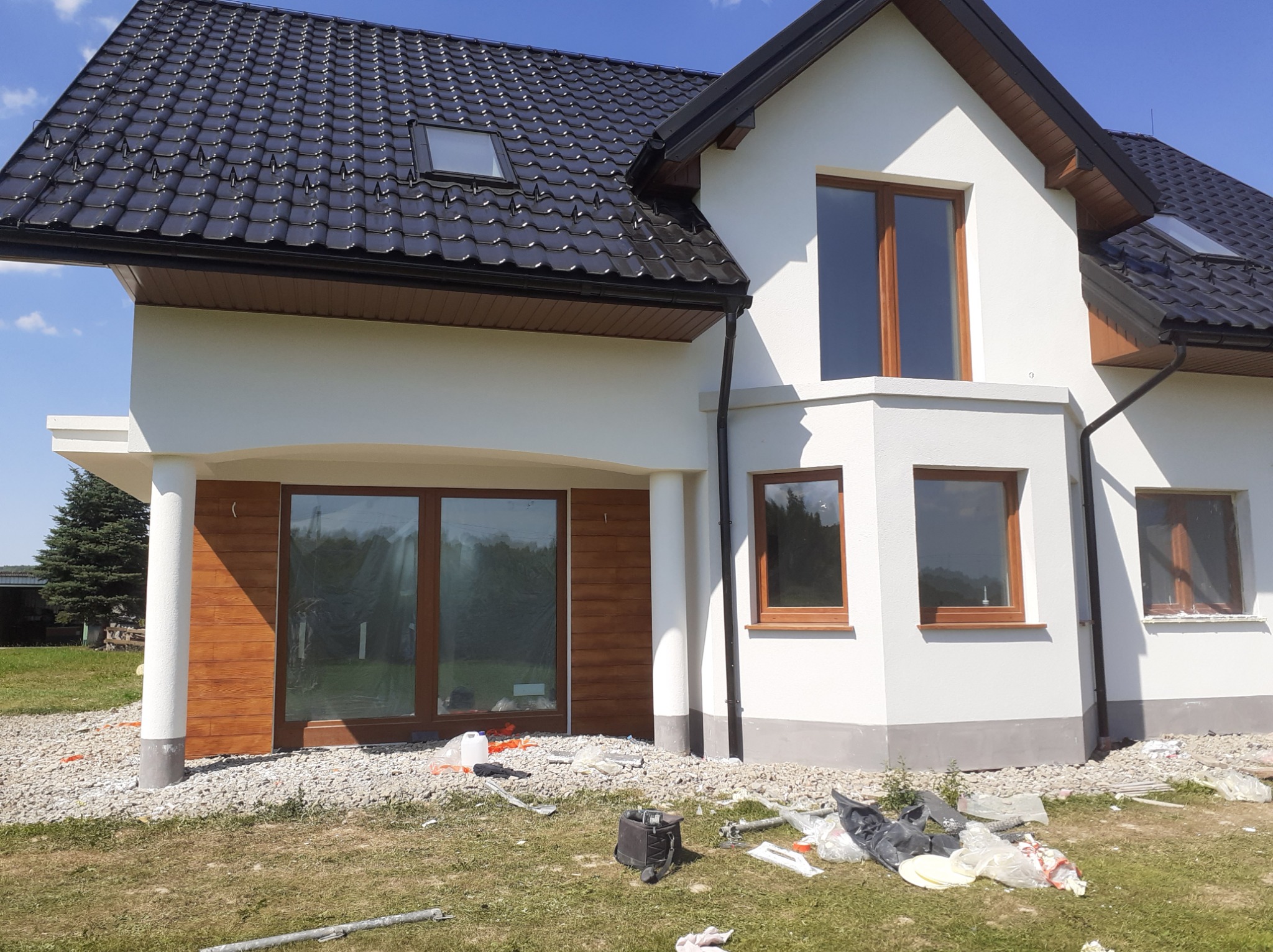 Biały dom jednorodzinny z ciemną dachówką i drewnianymi oknami. Widoczne wykończenia elewacji i otoczenie w trakcie prac. Ujęcie z poziomu gruntu.