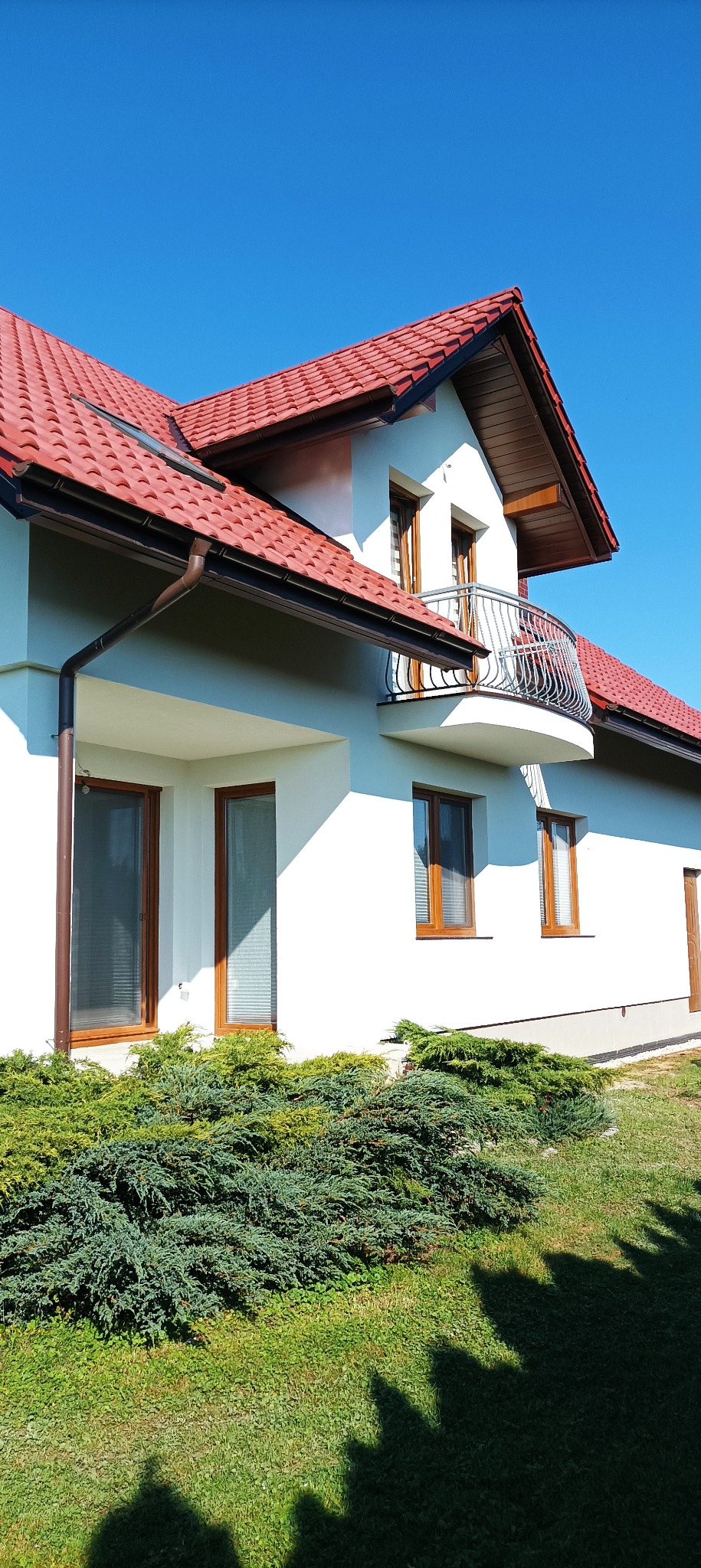 Biały dom jednorodzinny z czerwonym dachem i balkonem z metalową balustradą, widoczny z poziomu gruntu, w słoneczny dzień. Krzewy przed budynkiem.