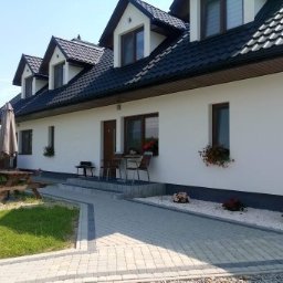 EDIT-BUD Edyta Gadzina - Biały dom z czarnym dachem, widok z zewnątrz. Uwagę przykuwają kwiaty w donicach pod oknami i starannie ułożony chodnik z kostki brukowej.