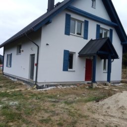 EDIT-BUD Edyta Gadzina - Biały dom z niebieskimi okiennicami i czerwoną stolarką drzwiową, świeżo po pracach elewacyjnych. Widoczne wykończenia wokół budynku i stos piasku.
