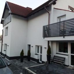 EDIT-BUD Edyta Gadzina - Biała elewacja domu z ciemną podmurówką, balkonem z poziomymi barierkami i brukowanym podjazdem. Widoczne detale architektoniczne i zadbana zieleń.