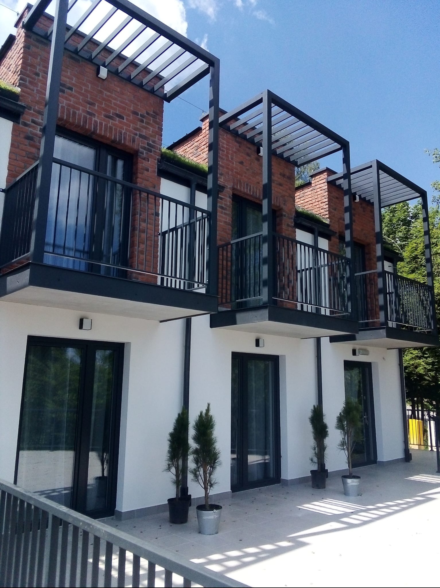 Nowoczesna elewacja budynku z cegły i białego tynku, balkony z czarną metalową konstrukcją i balustradami. U dołu małe drzewka w donicach.