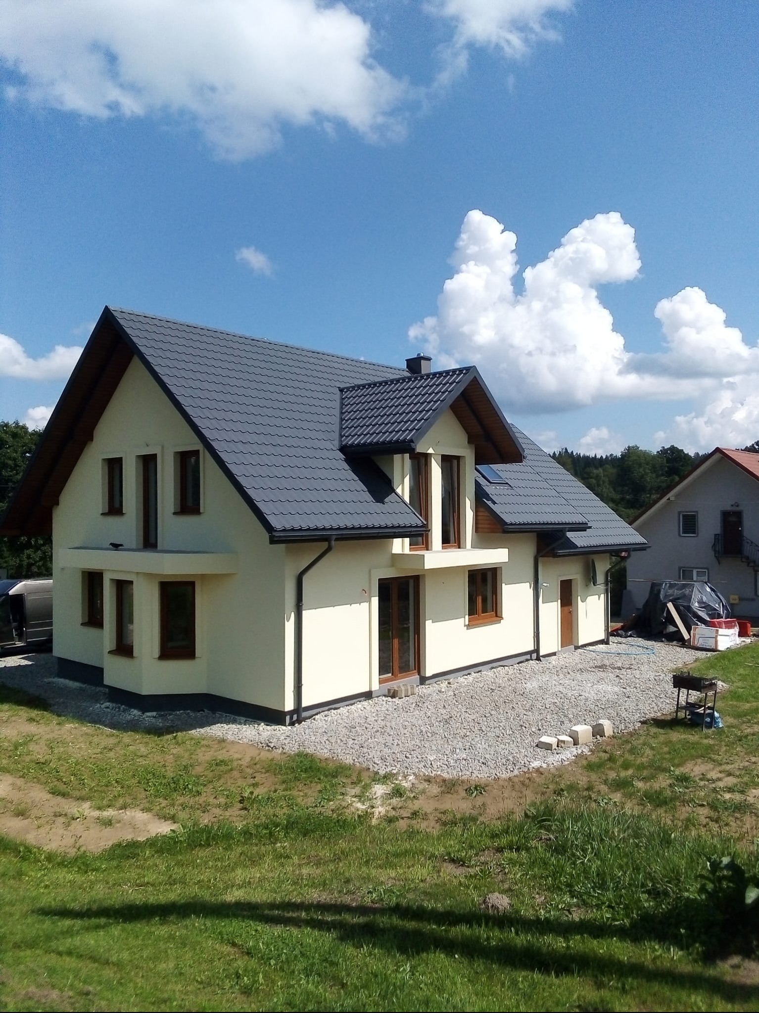 Jasny, dwukondygnacyjny dom z ciemnoszarą dachówką, brązowymi oknami i kremową elewacją, otoczony żwirem i zielenią, pod błękitnym niebem z obłokami.