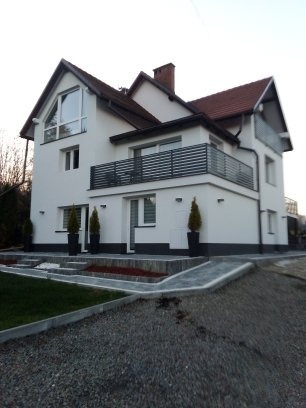 Biały dom jednorodzinny z balkonem i nowoczesnym ogrodzeniem, widok z poziomu gruntu. Uwagę zwracają małe drzewka w donicach przy wejściu i szara kostka brukowa.