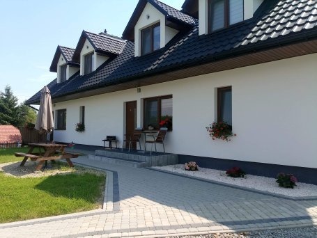 Biały dom z czarnym dachem, widok z zewnątrz. Uwagę przykuwają kwiaty w donicach pod oknami i starannie ułożony chodnik z kostki brukowej.