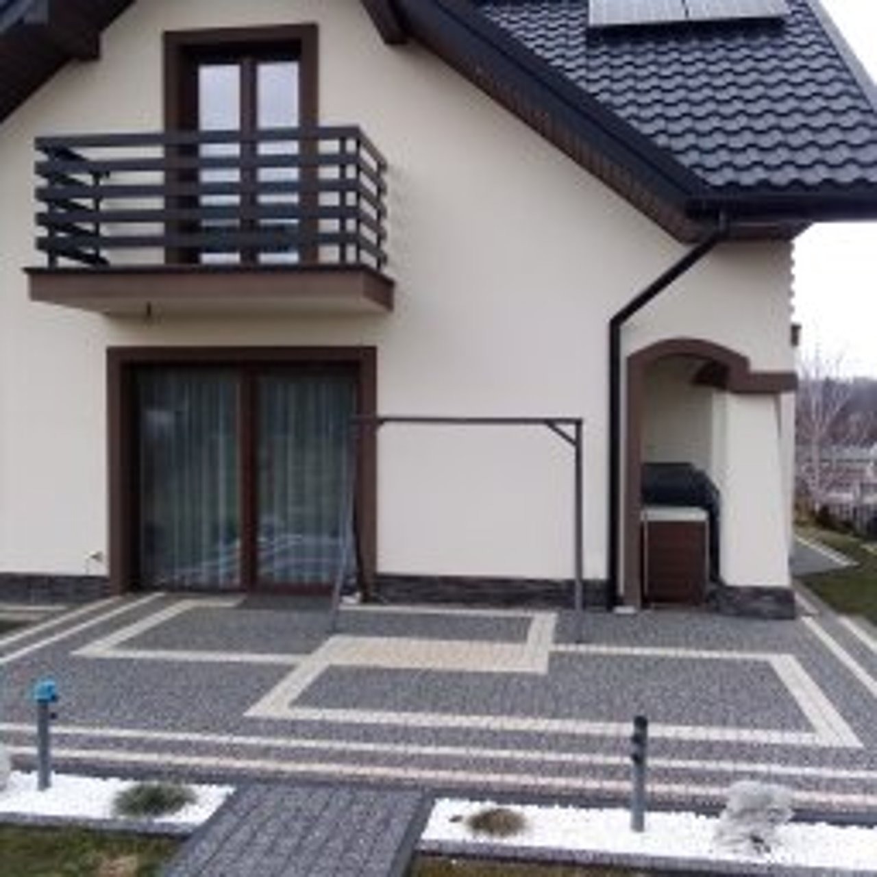 Elewacja domu jednorodzinnego z balkonem i ozdobnym wykończeniem w Ustrzykach Dolnych. Jasna kolorystyka ścian, ciemna stolarka okienna i dachówka.