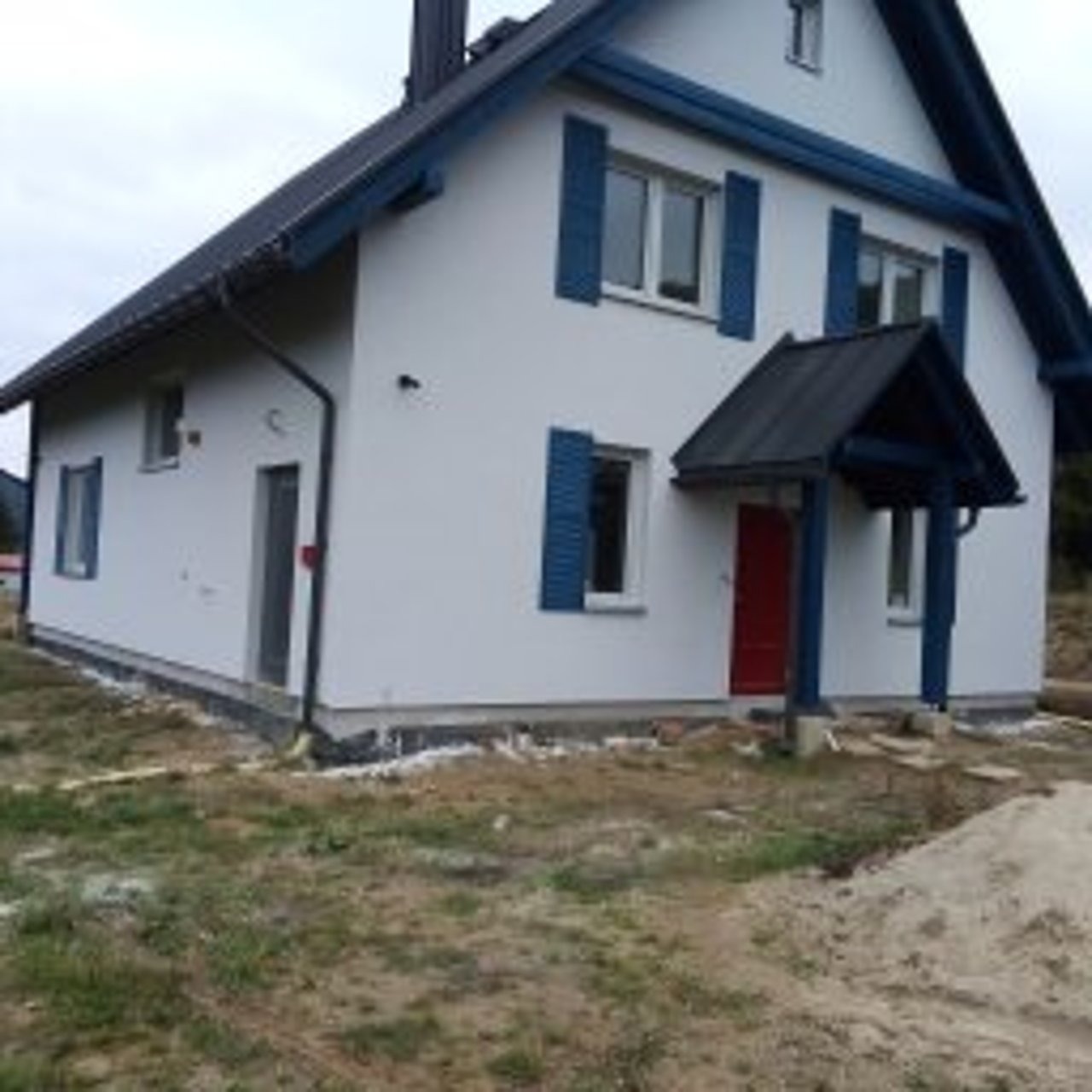 Biały dom z niebieskimi okiennicami i czerwoną stolarką drzwiową, świeżo po pracach elewacyjnych. Widoczne wykończenia wokół budynku i stos piasku.