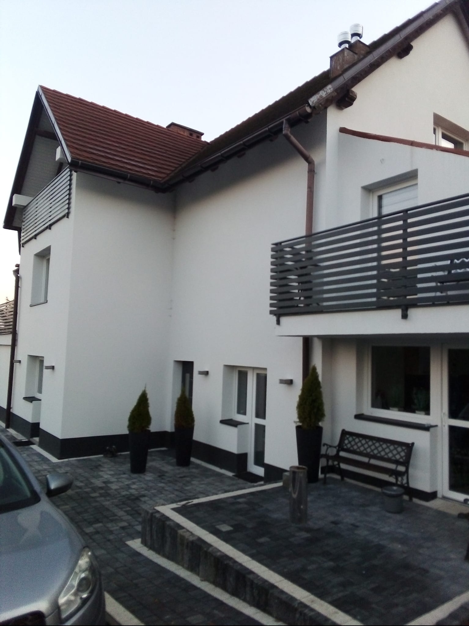 Biała elewacja domu z ciemną podmurówką, balkonem z poziomymi barierkami i brukowanym podjazdem. Widoczne detale architektoniczne i zadbana zieleń.