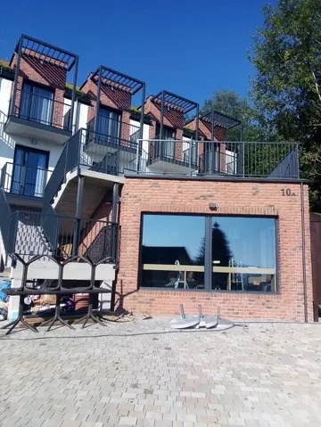 Nowoczesny budynek z ceglaną elewacją, balkonami z metalowymi konstrukcjami i zewnętrznymi schodami. Widoczne duże okno i numer budynku '10a'.