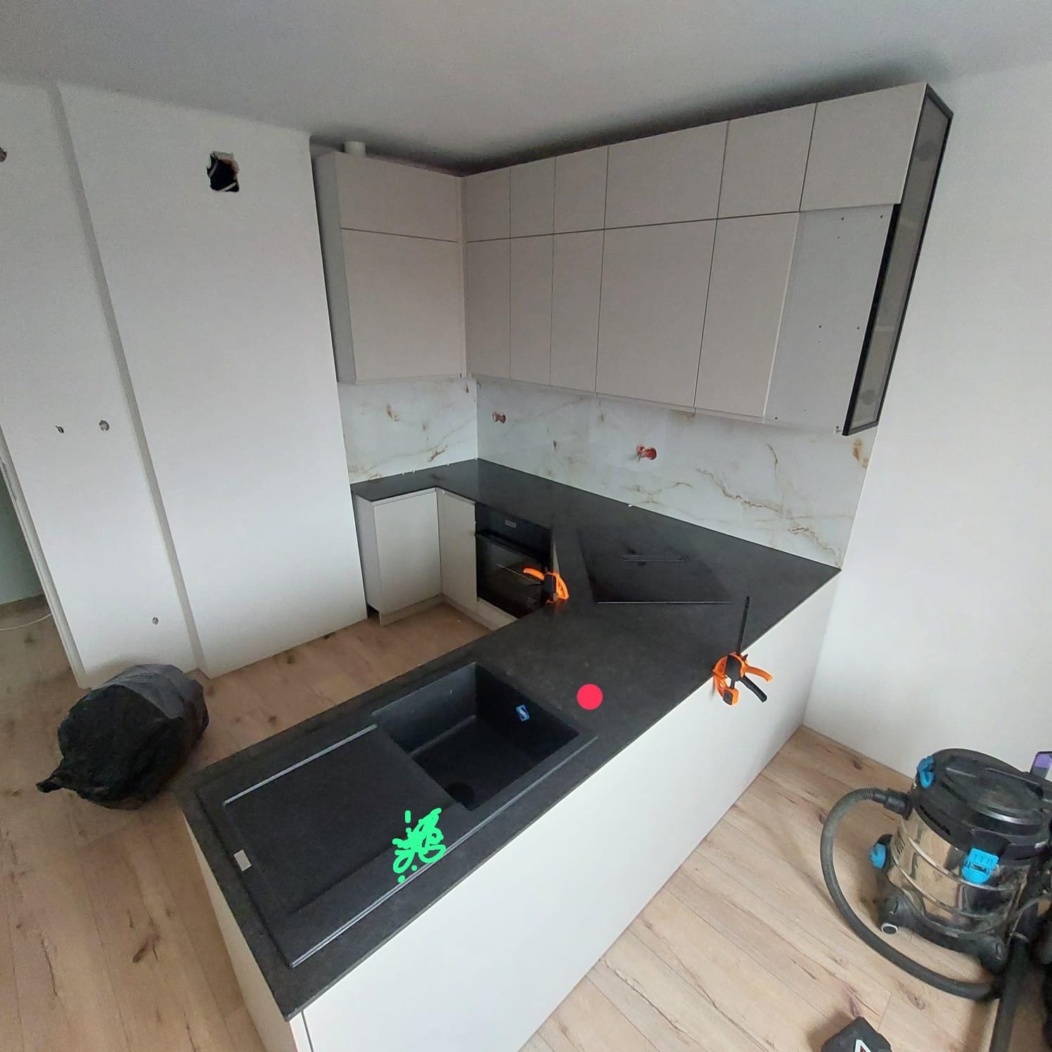 Montaż białej, nowoczesnej kuchni z czarnym blatem i marmurowym backsplashiem. Widoczne narzędzia i odkurzacz. Etap prac wykończeniowych, surowy stan ścian.