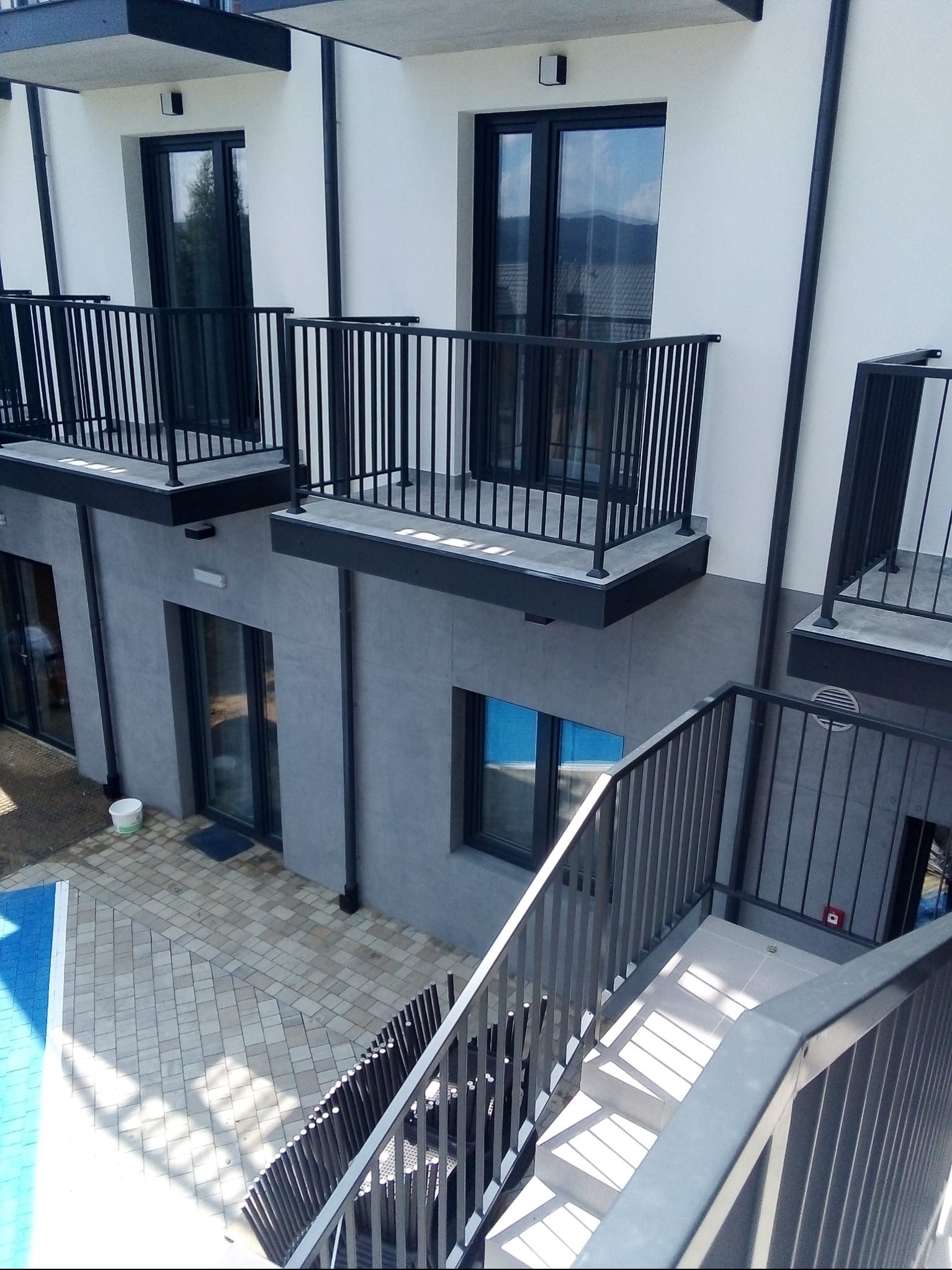 Nowoczesny budynek z balkonami o czarnych balustradach i szarą elewacją. Widok z góry na schody i fragment basenu. Minimalistyczny design.