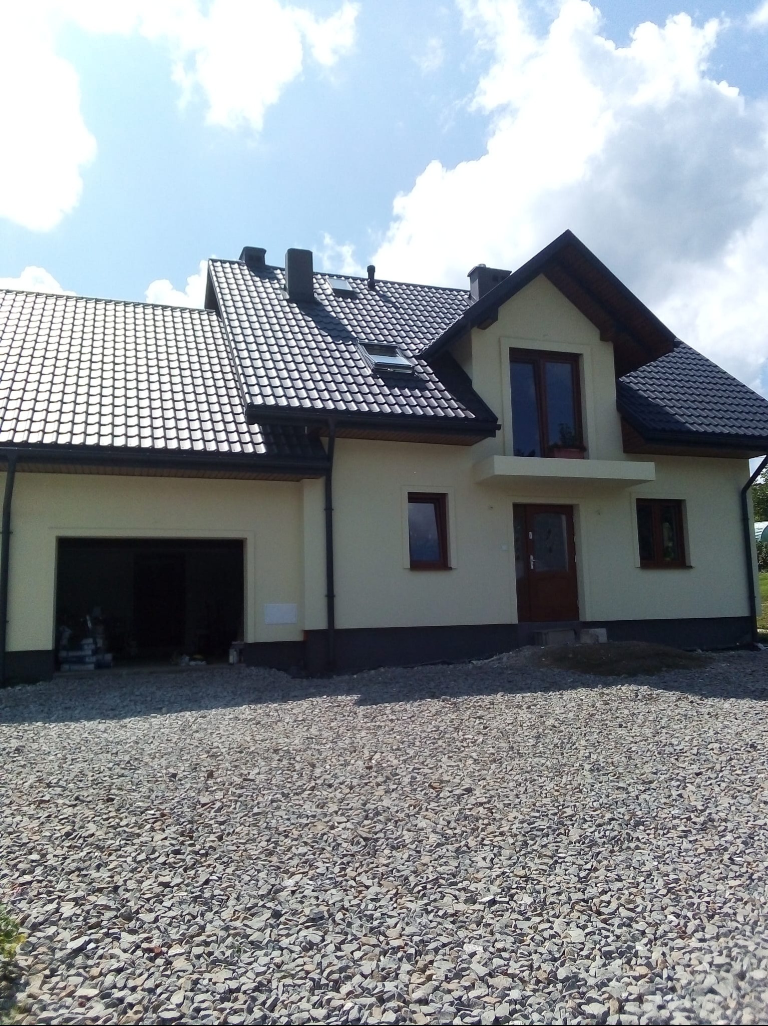 Jasny dom jednorodzinny z ciemnym dachem i garażem, otoczony kamiennym podłożem. Widoczne okna i drzwi wejściowe. Błękitne niebo z chmurami w tle.