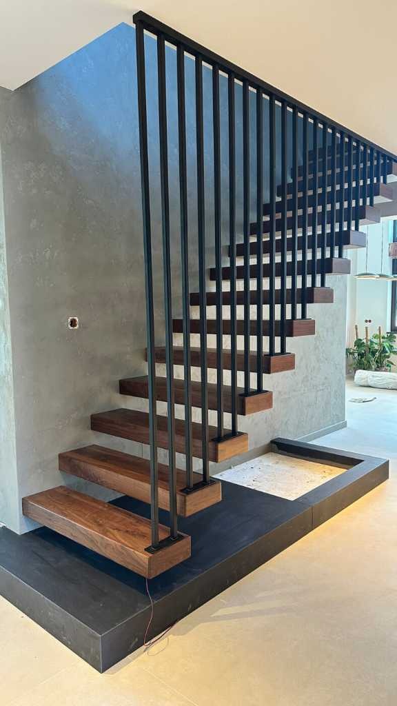 Nowoczesne schody z drewnianymi stopniami i czarną metalową balustradą na tle szarej ściany z widocznym gniazdkiem elektrycznym. Minimalistyczny design.