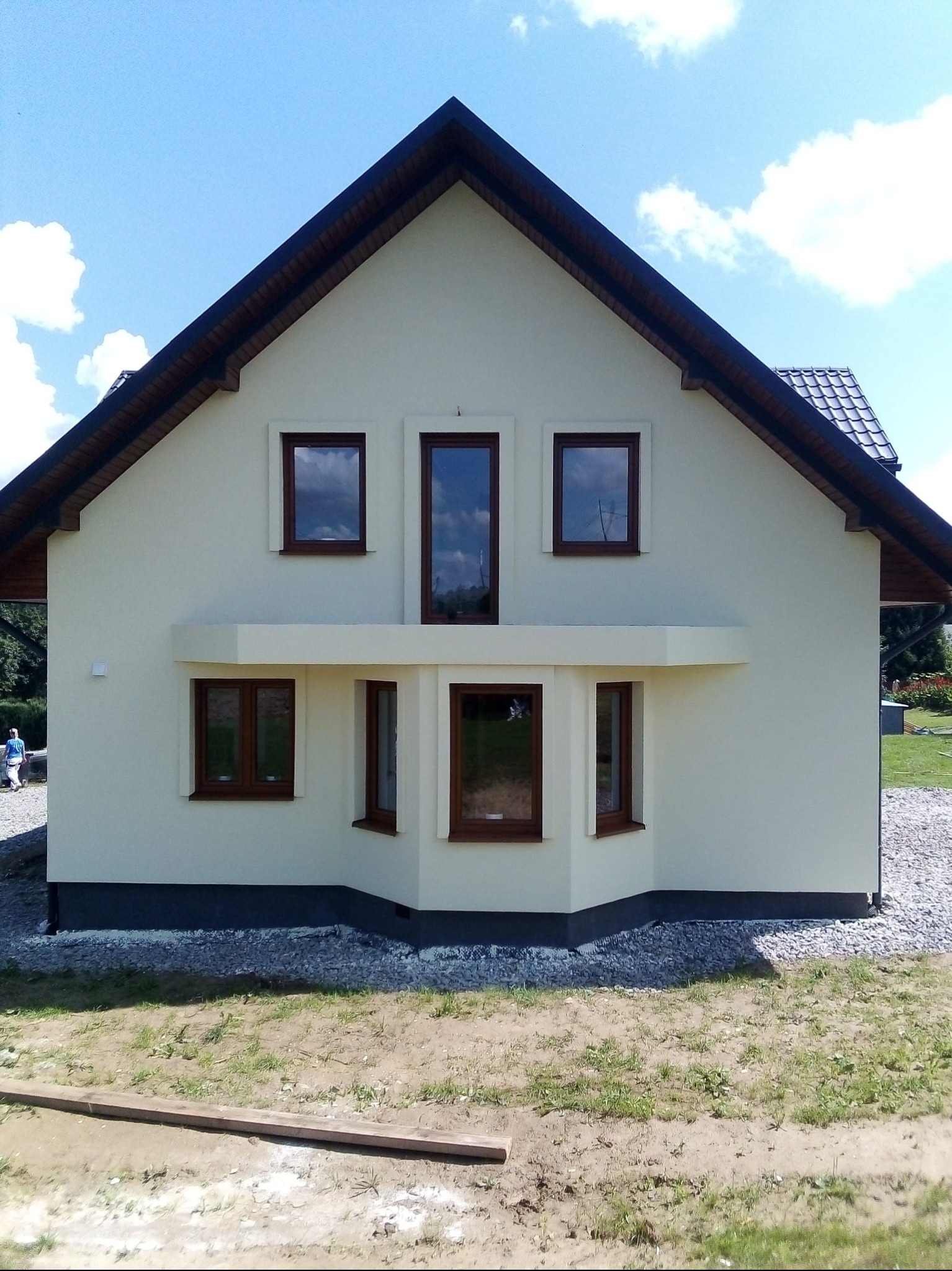 Jasna elewacja domu jednorodzinnego z ciemną podmurówką i brązowymi oknami, widok z przodu w słoneczny dzień. Charakterystyczny wykusz z oknami na parterze.