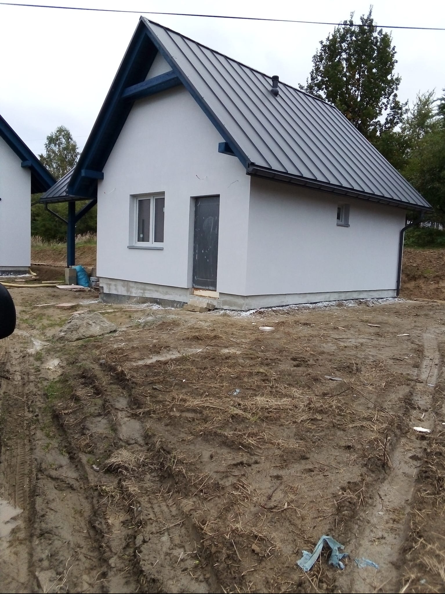 Biały budynek z szarym dachem i niebieskimi elementami wykończeniowymi, widoczny z boku na tle ziemi i zieleni. Małe okno i ciemne drzwi.