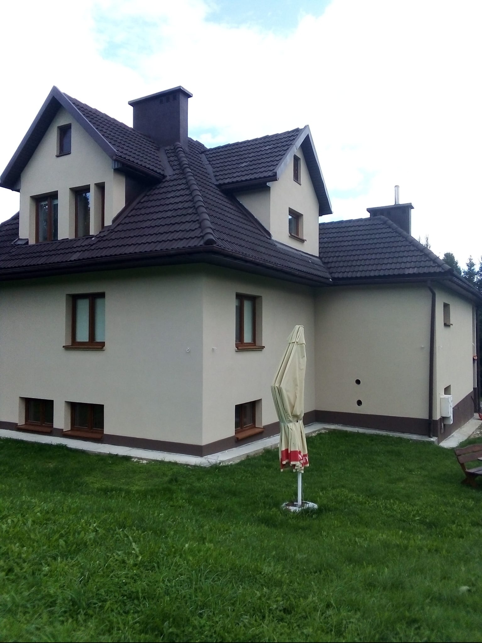 Dom jednorodzinny z beżową elewacją, brązowym dachem i stolarką okienną. Widoczny trawnik i częściowo złożony parasol ogrodowy. Ujęcie z poziomu gruntu.