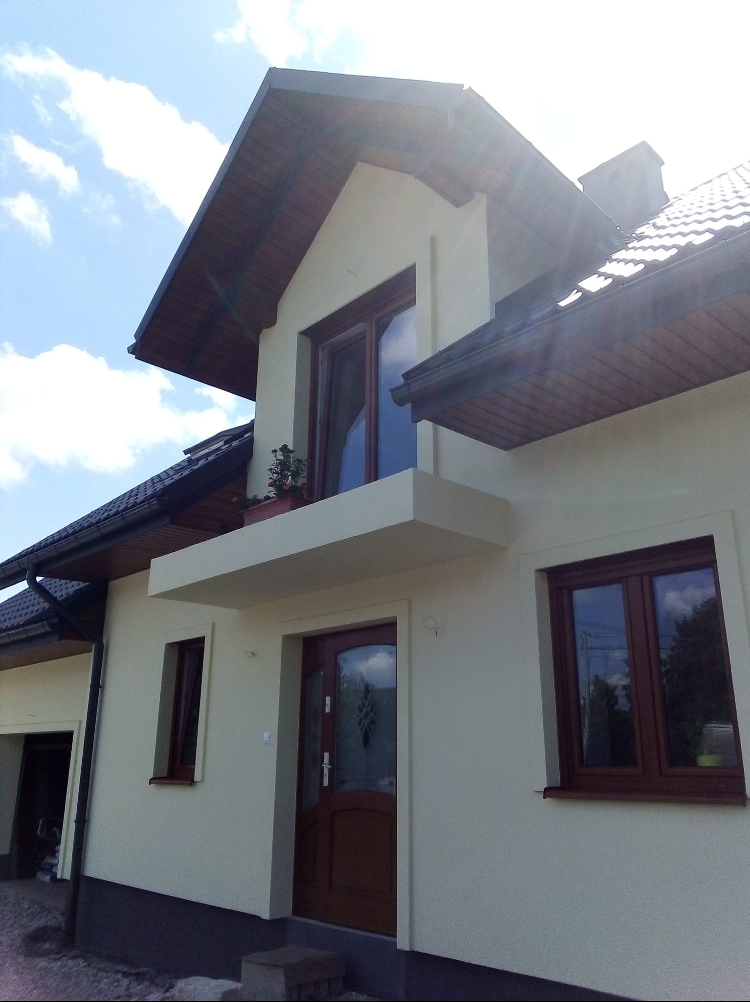 Jasna elewacja domu jednorodzinnego z brązowymi oknami i drzwiami, balkon z kwiatami, dachówka i błękitne niebo w tle. Ujęcie z dołu, podkreślające bryłę budynku.