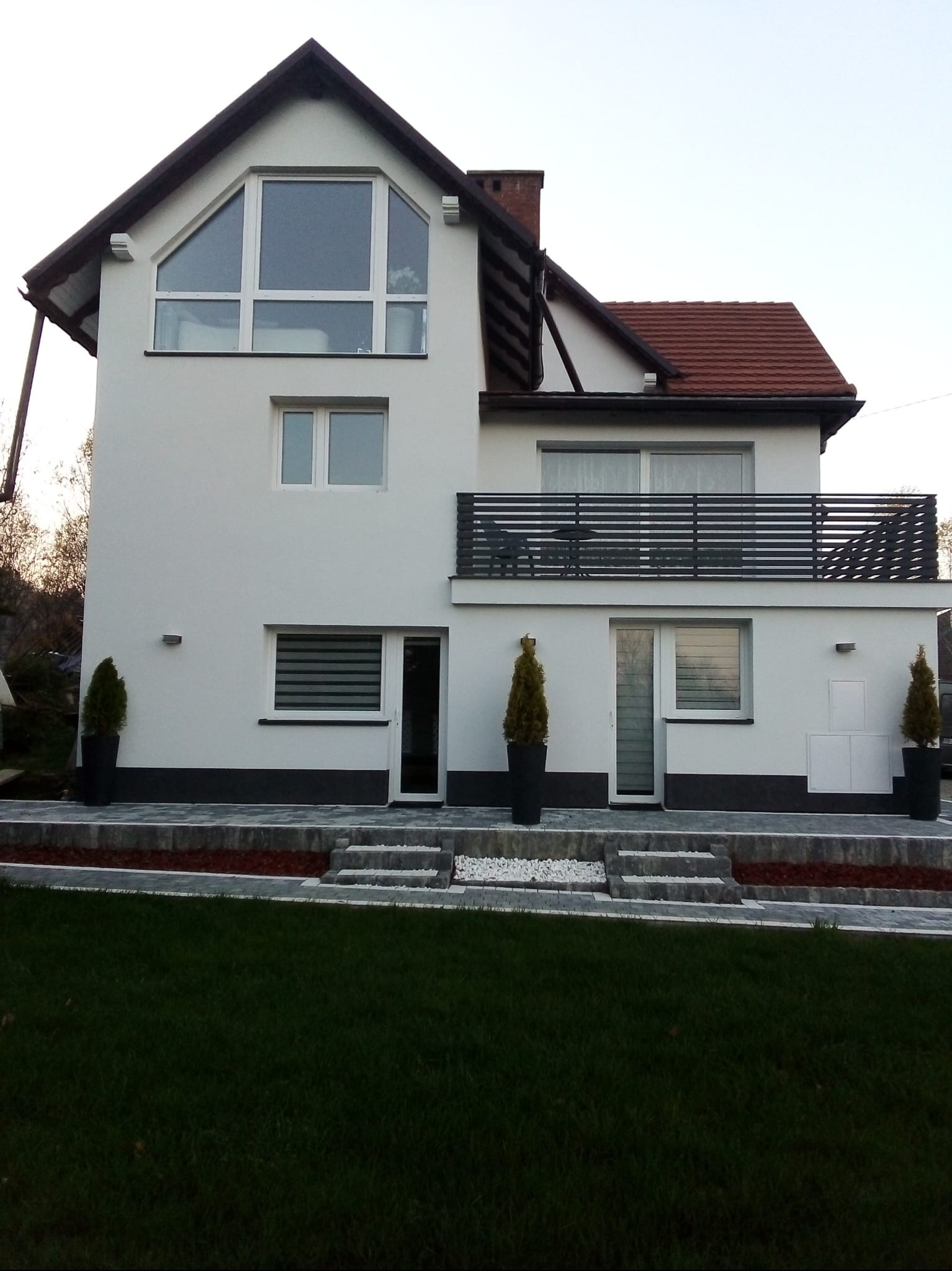 Biała elewacja dwukondygnacyjnego domu z balkonem i dużym oknem trójkątnym na poddaszu. Uwagę przykuwają geometryczne detale i minimalistyczny styl.