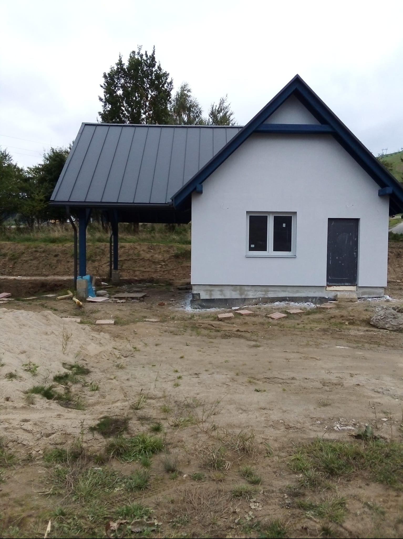 Biały budynek z szarym dachem i niebieskimi elementami wykończeniowymi, w trakcie budowy, z widocznym terenem wokół i fragmentem zadaszenia.