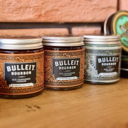 Sylvan Barbershop - Trzy słoiczki pomady do włosów Bulleit Bourbon na drewnianym blacie, z ceglaną ścianą w tle. Różne warianty produktu, widoczne detale etykiet.